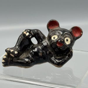 May include: A black ceramic bear figurine with red inner ears, white eyes, and white accents on its paws. The bear is lying down with its arms and legs outstretched, giving it a relaxed appearance. The figurine has a glossy finish.