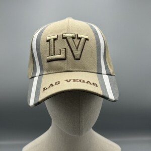 Pode incluir: Um boné de beisebol bege com listras cinzas e brancas. O boné tem as letras "LV" bordadas em marrom na frente e as palavras "LAS VEGAS" bordadas em marrom na aba.
