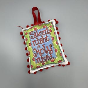 May include: A Christmas ornament with the words "Silent night Holy night" on a white square with a green border and red polka dots. The ornament has a red ribbon for hanging and is trimmed with red pom-poms.