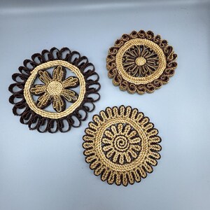 May include: Three round woven coasters with floral designs. The coasters are made from light and dark brown natural fibres. Each coaster has a unique pattern, with one featuring a flower shape in the centre.
