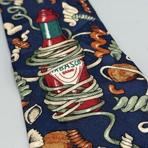 May include: A navy blue necktie featuring a red Tabasco sauce bottle wrapped in green and white pasta shapes. The tie has a repeating pattern of pasta and sauce bottles.