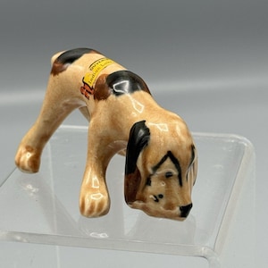 May include: A ceramic figurine of a dog in a downward pose, with a tan body, black patches, and a brown ear. The dog has a small yellow tag that reads "Souvenir Indian Lake". The figurine is on a clear surface.