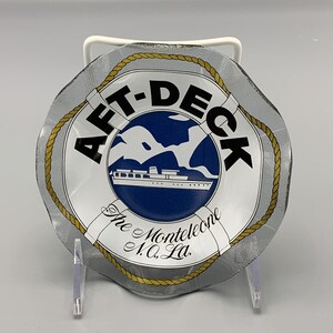 以下が含まれることがあります： 銀と金のデザインが施された白いガラス製の灰皿。灰皿は、黒い文字で「Aft-Deck The Montelecione N.O. La.」と書かれた救命浮き輪のデザインです。