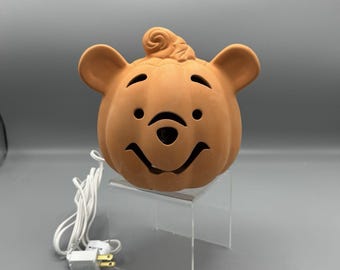 May include: A light brown ceramic pumpkin-shaped night light with a bear face design. The light has a curly stem and a smiling face with black eyes. It includes a white electrical cord and plug.