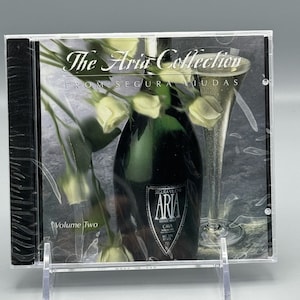 May include: A CD case with a white background featuring a bottle of sparkling wine with a white label that reads "SEQUIRA VILAS ARIA ESTATE BOTTLED CAVA ALC 12% BY VOL 750". The bottle is surrounded by white roses and a glass of sparkling wine. The text "The Aria Collection FROM SEGURA VIUDAS Volume Two" is printed on the case.