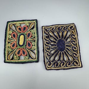 May include: Two rectangular coasters with detailed woven patterns. One has a central oval with red, yellow, and green accents, the other a blue centre with swirling gold designs. Both have dark blue borders.