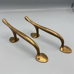 Może przedstawiać: Para antycznych mosiężnych klamki drzwiowych o zakrzywionym kształcie. Klamki wykonane są z litego mosiądzu i mają polerowane wykończenie.