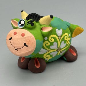 May include: A green ceramic cow figurine with a winking eye, brown hooves, and a yellow tail. The cow is decorated with white and yellow flowers and glitter.