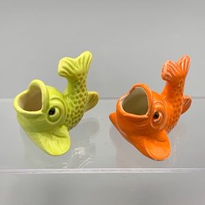 Może przedstawiać: Dwa ceramiczne doniczki w kształcie ryb, jedna zielona, a druga pomarańczowa, z otwartymi ustami i uniesionymi ogonami. Obie są małe i mają fantazyjny wygląd.