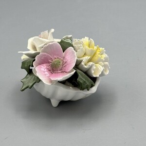 May include: A white porcelain floral arrangement. It features a pink flower with a green center, white and yellow flowers, and green leaves. The flowers are in a small, white, scalloped bowl with three small feet.