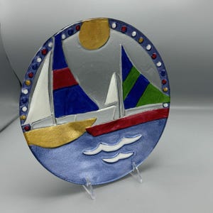 Puede incluir: Un plato de vidrio azul con un diseño en relieve de dos veleros en un mar azul con olas blancas. Los veleros están pintados en rojo, azul, verde y blanco. El plato tiene un borde dorado con puntos rojos y blancos.
