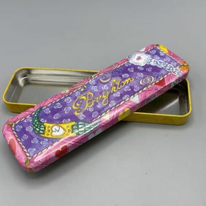 May include: A rectangular metal pencil case with a yellow interior and a decorative exterior. The exterior features a purple background with the word "Brighton" in gold script, along with floral and heart designs in pink and white.