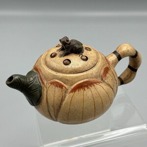 May include: A small, ceramic teapot shaped like a lotus flower with a frog on top. The teapot is decorated with brown and orange accents.
