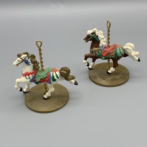May include: Two miniature carousel horses, one white with brown accents and one brown with gold accents, both with colorful saddles and a gold pole.