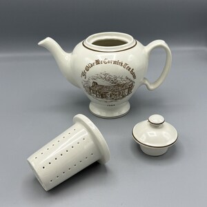 Può includere: Teiera in ceramica bianca con un design marrone che recita "Ye Olde McCormick Tea Room" e una data del 1983. La teiera è dotata di un filtro e di un coperchio.