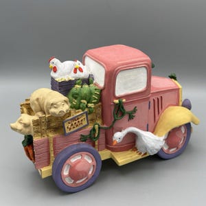May include: A pink and purple farm truck figurine with a load of farm animals, including pigs, chickens, and a goose. The truck bed has a sign that reads "The Carrot Farm."