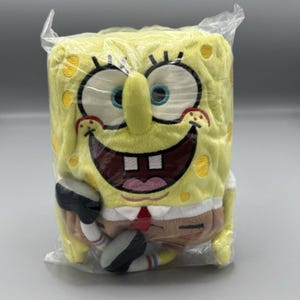 Puede incluir: Un peluche amarillo de Bob Esponja, empaquetado en plástico transparente. El juguete tiene una gran cara sonriente, pestañas negras y una corbata roja. El juguete rectangular tiene lunares amarillos.