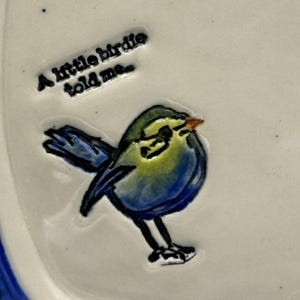 Könnte beinhalten: Keramikplatte mit einem erhabenen Design eines blauen und gelben Vogels. Der Vogel sitzt auf einer weißen Oberfläche, darüber der Text "A little birdie told me...". Die Platte hat einen blauen Rand.