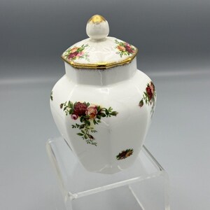 Peut inclure: Un pot en céramique blanche avec un couvercle doré et un motif floral. Le pot a une forme hexagonale et est décoré de roses rouges, roses et vertes.