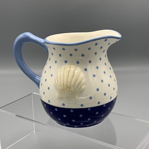 May include: A white ceramic pitcher with a blue rim and blue polka dots. The pitcher has a blue handle and a raised seashell design. The bottom half of the pitcher is blue with white polka dots.