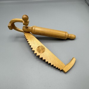 May include: A light-colored wooden cheese slicer with a saw-like blade and a rolling pin handle. The blade has a starburst design and the handle has text. The slicer is made of wood.