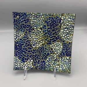 May include: A square ceramic dish with a blue, white, and gold mosaic pattern. The dish is raised on three clear acrylic stands.