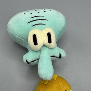 May include: A soft toy of the cartoon character Squidward Tentacles. The toy has a light blue head with three black lines, large cream-coloured eyes, and a long nose. He is wearing a yellow shirt.