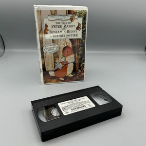 Può includere: Un nastro VHS della storia per bambini "La storia di Peter Rabbit e Benjamin Bunny" di Beatrix Potter. Il nastro è in una custodia nera con il titolo e l'autore stampati sull'etichetta. La copertina presenta un coniglio dei cartoni animati che indossa una giacca blu.