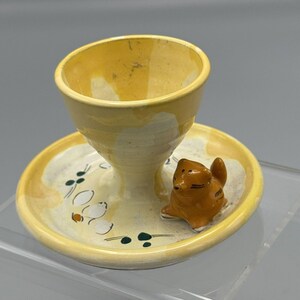 May include: A yellow ceramic egg cup with a matching saucer. The saucer has a small orange cat figurine on it and a white and green floral design.