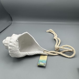 May include: White ceramic seashell-shaped hanging planter with a textured surface. The planter is suspended by a natural rope. A tag with the words "Hanging Shell" is attached to the rope.