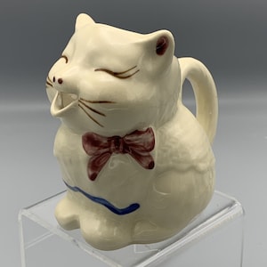 Peut inclure: Un pot à crème en céramique blanche en forme de chat avec un nœud papillon brun et des accents bleus. Le chat a la bouche grande ouverte et une expression espiègle.