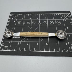 May include: A double-ended melon baller with a wooden handle and stainless steel scoops. The handle is approximately 4 inches long. The scoops are silver-colored and attached to the handle with brass-colored rivets. The tool is designed for scooping melon and other fruits.