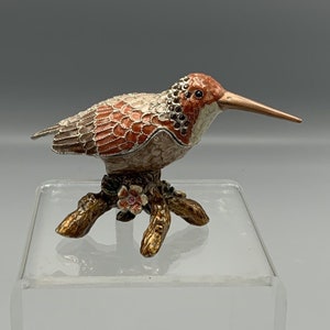 Pode incluir: Uma pequena figura de pássaro de esmalte, detalhada com cuidado, com um bico longo, pousada em um galho com uma flor rosa. O pássaro é pintado em tons de marrom, laranja e branco, com detalhes prateados.