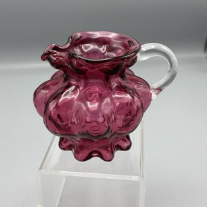 May include: A small, antique, cranberry-colored glass pitcher with a clear glass handle. The pitcher has a ruffled rim and a bulbous body with a textured, ribbed design. The handle is curved and attached to the side of the pitcher.