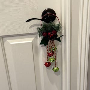 May include: A Christmas door hanger featuring a dark green and red color scheme. The door decoration includes faux pine needles, red berries, and hanging ornaments in red and green. The hanger is attached to a black door handle.