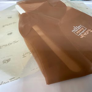 Könnte beinhalten: Ein Paar transparente, braune Nylonstrümpfe, gefaltet und auf Seidenpapier liegend. Die Strümpfe sind nahtlos und tragen die Aufschrift "100% Nylon" und "Seamless". Das Seidenpapier hat ein sich wiederholendes Muster aus Text und kleinen Illustrationen.