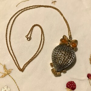May include: A gold-toned necklace with an owl pendant. The owl pendant features a silver cage-like body, gold-toned accents, and two orange-colored eyes. The necklace chain is delicate and gold-toned.