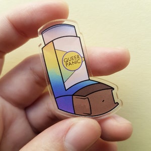 Puede incluir: Un llavero de plástico transparente con forma de inhalador con un diseño degradado arcoíris y el texto "QUEER PANIC" en un círculo amarillo.