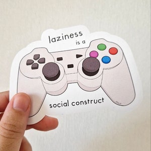 May include: White sticker with a cartoon illustration of a video game controller. The text on the sticker reads "Laziness is a social construct".
