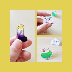 Può includere: Tre spille in acrilico con design diversi. La prima spilla è un inalatore viola e giallo con il testo "Non Binary Panic". La seconda spilla è un controller per videogiochi bianco con il testo "Laziness is social commitment". La terza spilla è un dessert verde e rosso con il testo "There's no guilt in pleasure".