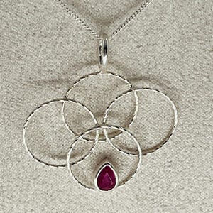 May include: A silver pendant necklace with a design of four interlinked circles. The pendant has a textured finish and a teardrop-shaped, deep red gemstone at the centre. The chain is a fine silver cable.