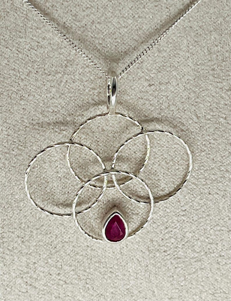 May include: A silver pendant necklace featuring a cluster of four interconnected circles. The pendant has a textured design and a teardrop-shaped, deep red gemstone at the center. The chain is a delicate silver cable.