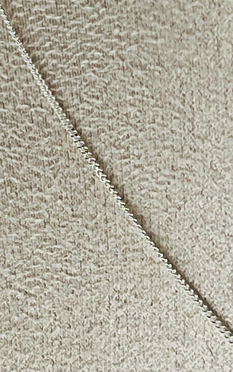 May include: A close-up of a delicate silver chain with a textured, woven design. The chain is positioned diagonally against a neutral, textured background, creating a contrast in textures and tones. The chain's intricate details are highlighted.