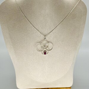 May include: A silver pendant necklace featuring a floral design with a small, teardrop-shaped, red gemstone at the center. The pendant is suspended from a delicate silver chain. The necklace is displayed on a mannequin.