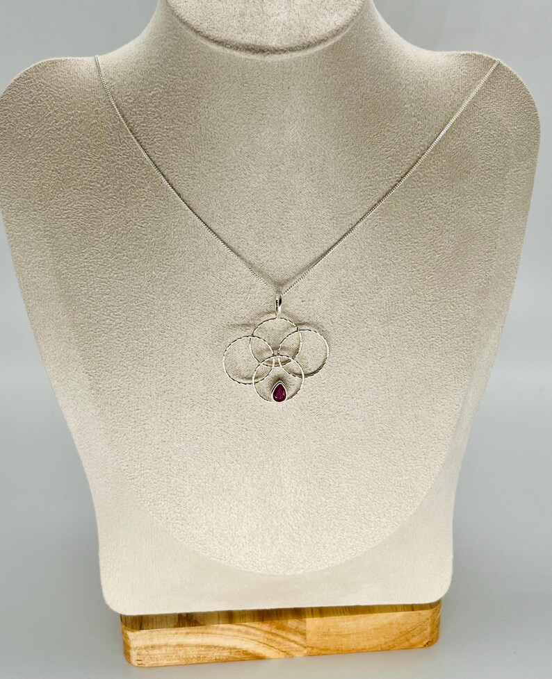 May include: A silver pendant necklace featuring a floral design with a small, teardrop-shaped, red gemstone at the center. The pendant is suspended from a delicate silver chain. The necklace is displayed on a mannequin.