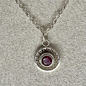 May include: A silver necklace with a round pendant featuring a purple gemstone surrounded by a decorative border.