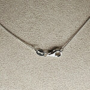 May include: Close-up of a silver chain necklace with a lobster clasp. The chain has a delicate, textured design. The clasp is open, showing the inner workings of the mechanism. The background is a neutral, textured surface.