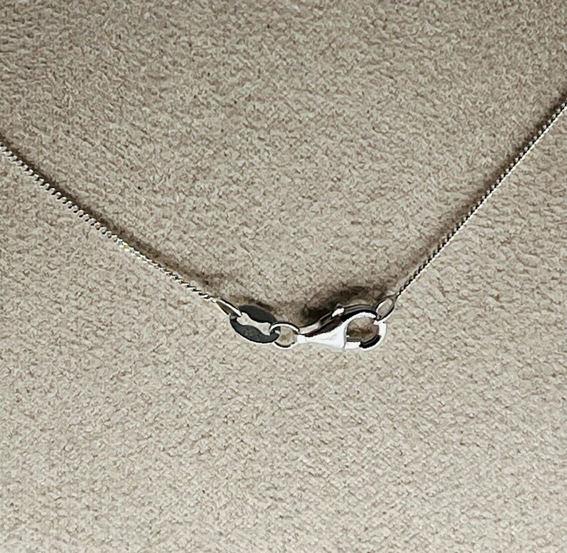 May include: Close-up of a silver chain necklace with a lobster clasp. The chain has a delicate, textured design. The clasp is open, showing the inner workings of the mechanism. The background is a neutral, textured surface.