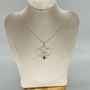 May include: A silver necklace with a pendant featuring four interconnected circles and a teardrop-shaped red gemstone. The necklace is displayed on a beige mannequin. The pendant design is delicate and intricate.