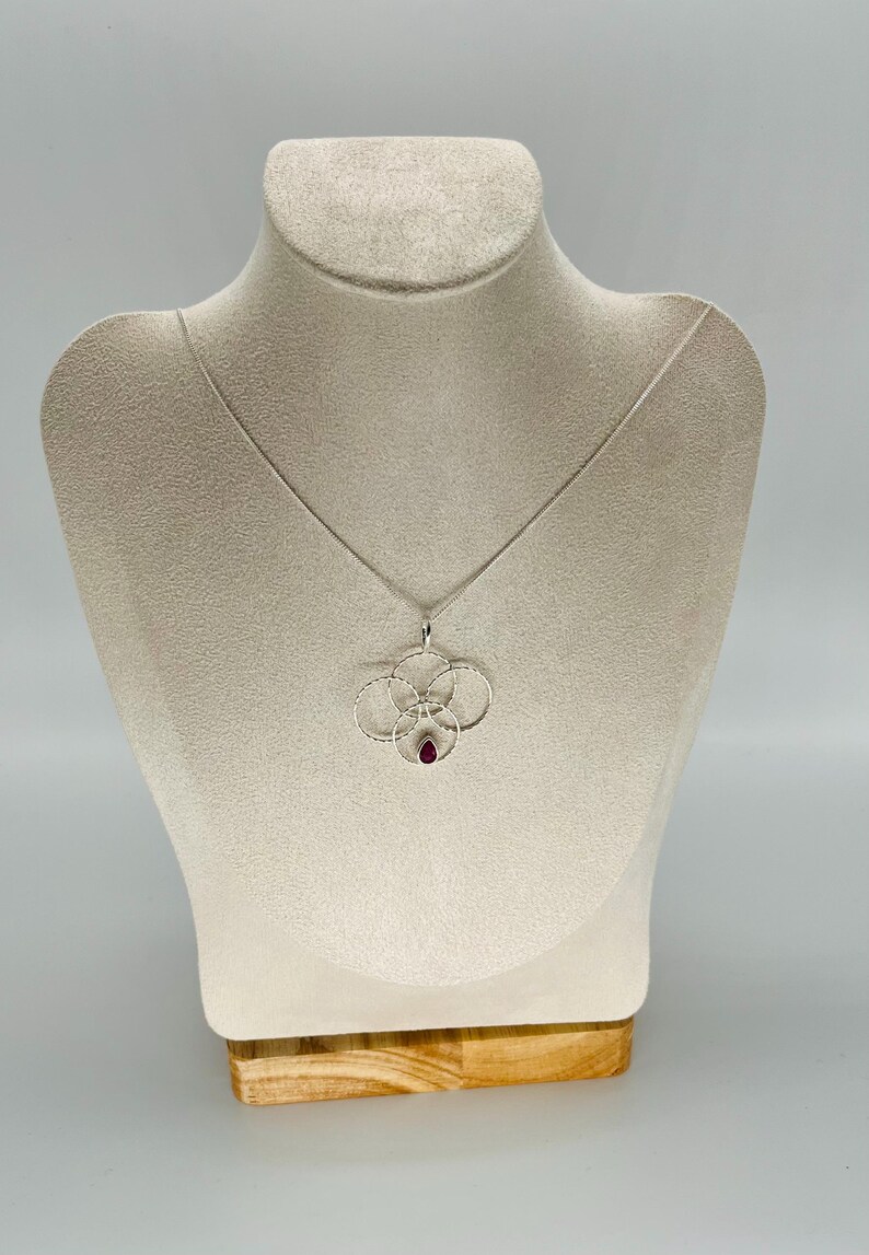 May include: A silver necklace with a pendant featuring four interconnected circles and a teardrop-shaped red gemstone. The necklace is displayed on a beige mannequin. The pendant design is delicate and intricate.